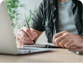Man writing on a notepad in front of a laptop.
