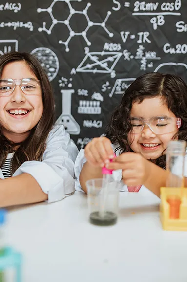Two girls smiling and playing with their STEM kit wearing lab safety gear