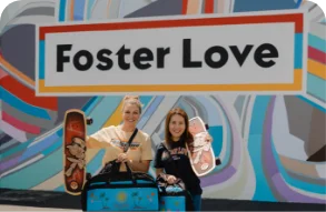 Two volunteers holding up their service project Sweet Cases and Skateboards in front of the Foster Love mural